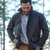 Man outdoors in a dark jacket, Monroe Poplin Shirt from Tom Beckbe, and khaki pants, hands in pockets, amidst blurred trees.