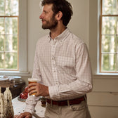A man in a Tom Beckbe Monroe Poplin Shirt holds a glass of beer in a sunny room with large windows.