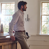 A man in a Monroe Poplin Shirt by Tom Beckbe leans against a wooden counter in a sunlit room with two large windows.