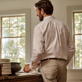 A man in a Tom Beckbe Monroe Poplin Shirt and beige pants stands by a kitchen counter holding a mug by windows.