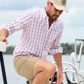 Man in a Tom Beckbe Monroe Poplin Shirt and cap steps onto a boat, with water and trees in the background.