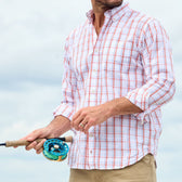 A man in a Tom Beckbe Monroe Poplin Shirt and khaki pants holds a fishing rod against a cloudy sky backdrop.
