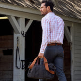 Man in a plaid Monroe Poplin Shirt and gray pants, by Tom Beckbe, walking away with a duffel bag with tan straps.