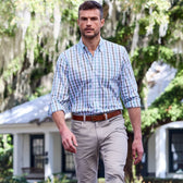 A man in a Monroe Poplin Shirt by Tom Beckbe, beige pants, walks outdoors with trees and a house behind him.