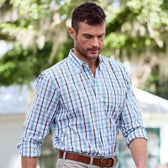 A man strolls outdoors in a Monroe Poplin Shirt by Tom Beckbe and khaki pants, with trees and a house in the background.