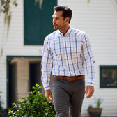 A man in a Monroe Poplin Shirt from Tom Beckbe and gray pants strolls by a white building with green shutters.