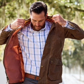 Man in a brown jacket adjusts the collar of his Tom Beckbe Monroe Poplin Shirt outdoors on a sunny day.