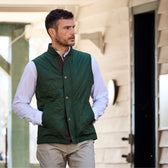 Man in a Monroe Poplin Shirt by Tom Beckbe, green quilted vest, beige pants, outdoors with hands in pockets.
