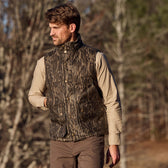 A man in a Tom Beckbe Fowler Vest (Mossy Oak Original Bottomland) and beige shirt stands outdoors with blurred trees behind.