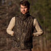 A man outdoors in a Tom Beckbe Fowler Vest, Mossy Oak Original Bottomland, beige shirt, hands in pockets among tall trees.