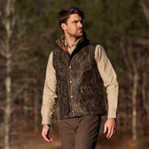 Man outdoors in Tom Beckbes Fowler Vest, Mossy Oak Original Bottomland, over beige shirt and brown pants with tree backdrop.