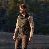 A man in a Tom Beckbe Kinsman Vest in Mossy Oak Original Bottomland and khaki shirt walks outdoors beneath sunny trees.