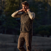Man wearing a Kinsman Vest in Mossy Oak Original Bottomland over a beige shirt, adjusting the collar outdoors.