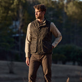 A man in a Tom Beckbe Kinsman Vest, Mossy Oak Original Bottomland, and beige shirt stands outdoors with trees behind.