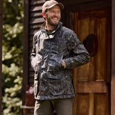 Man in a patterned jacket and cap smiles with hands in pockets outside a wooden building. Tom Beckbe Tensaw Jacket feels.