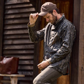 Man in a Tom Beckbe Tensaw Jacket in Mossy Oak Original Bottomland leans on a wooden building, adjusting his hat.