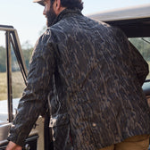 Wearing Tom Beckbes Tensaw Jacket in Mossy Oak Original Bottomland, a man steps into a car, blending with nature.
