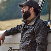 A man in a cap and a Tom Beckbe Tensaw Jacket (Mossy Oak Original Bottomland) leans against a white vehicle outdoors.