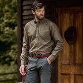 Man in a Tom Beckbe olive Culver Oxford Shirt and gray pants stands outside a wooden building, hands in pockets.