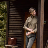 A man adjusts his Culver Oxford shirt cuff on a porch with a brown bag on a bench and wooden building in the background.