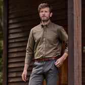A bearded man leans against a beam, wearing a breathable Tom Beckbe Culver Oxford Shirt outside a wooden building.
