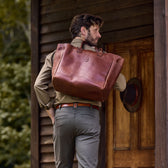 Person walking with a large brown leather tote, wearing a Tom Beckbe Culver Oxford Shirt, in front of a wooden building.