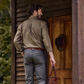 A man in a Tom Beckbe Culver Oxford Shirt and gray pants stands at a wooden door, holding a brown leather bag with a hat hanging.