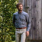 Man in a Tom Beckbe Culver Oxford Shirt and beige pants stands outside by a wooden structure among lush greenery.