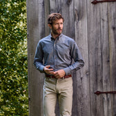 A man in a Tom Beckbe Culver Oxford Shirt stands by a rustic wooden wall with greenery in the background.