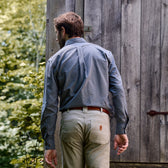 Man in Tom Beckbes Culver Oxford Shirt and beige pants strolls by a wooden structure among lush trees.