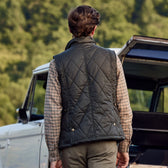 A person in a Tom Beckbe Paddock Quilted Vest stands by a vehicle amid trees, kept warm by HD Wool.