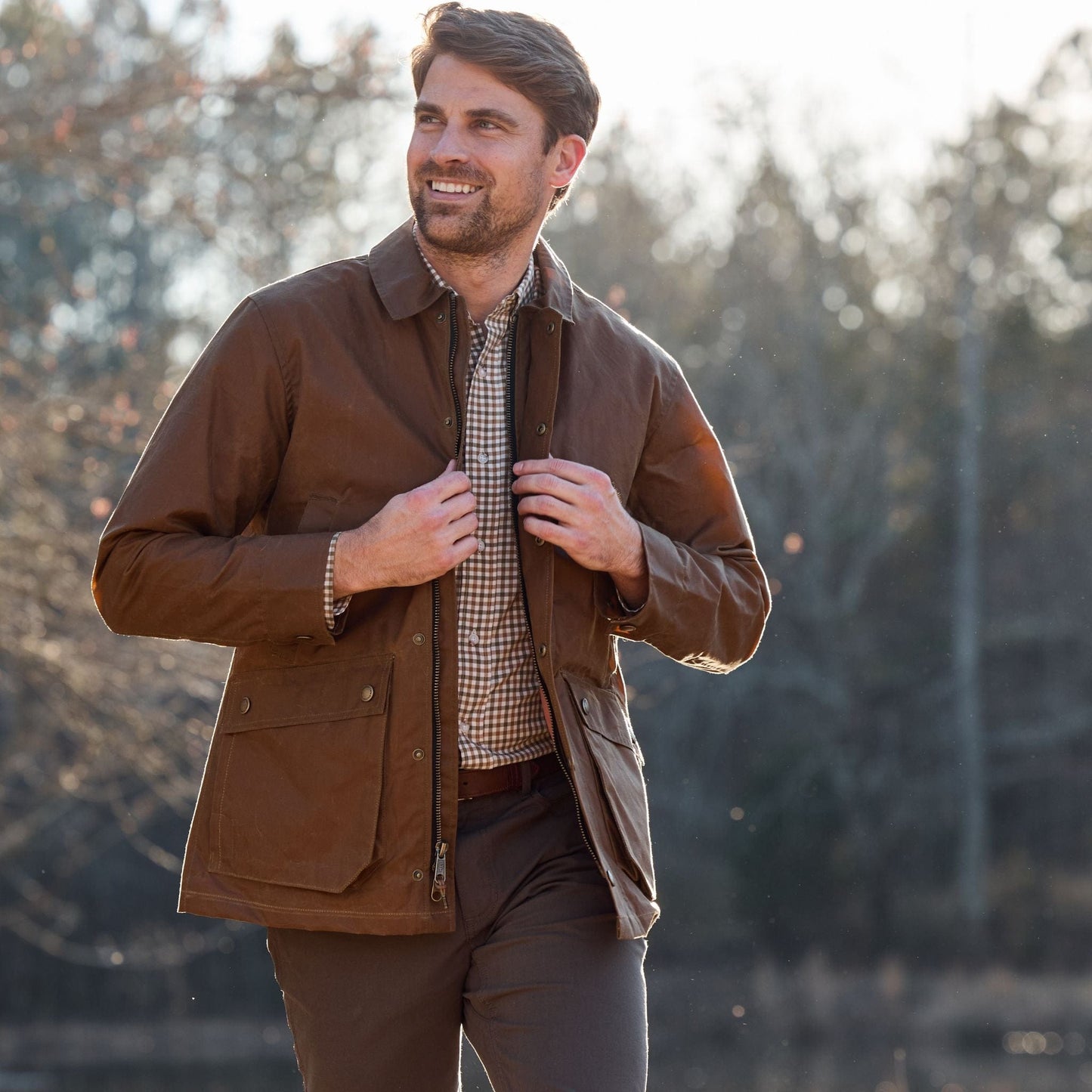 Man outdoors in a Tom Beckbe Piedmont Jacket over a plaid shirt, smiling with trees in the background.