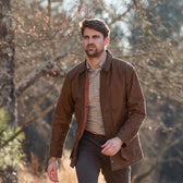A man in a Tom Beckbe Piedmont Jacket and plaid shirt walks outdoors in a wooded area during the day.