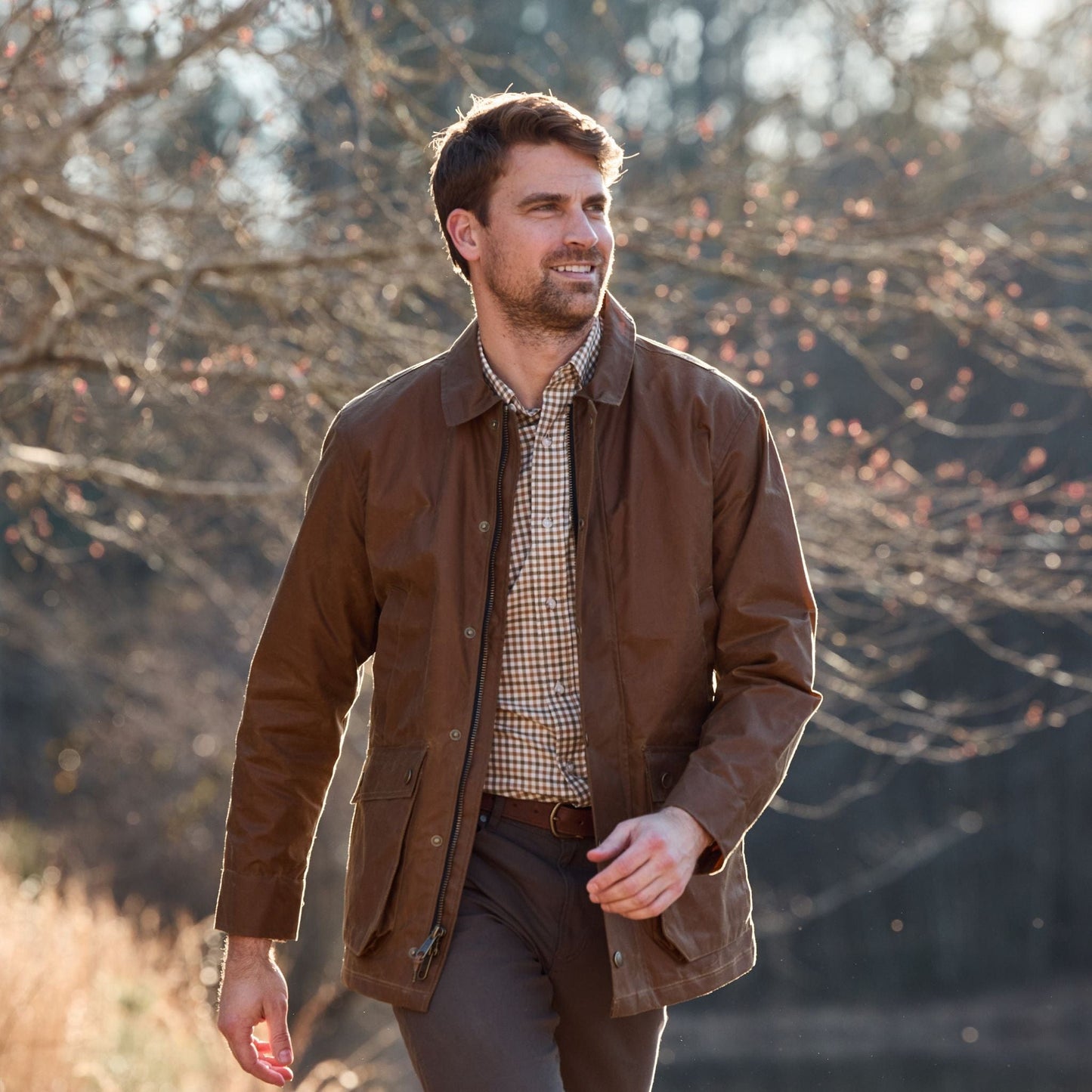 A man in a Tom Beckbe Piedmont Jacket and checked shirt walks outdoors on a sunny day with bare trees behind him.