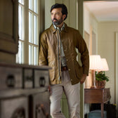 Man in a Tom Beckbe Piedmont Jacket and light pants stands by a window indoors, looking outside thoughtfully.