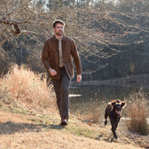 A man in a Tom Beckbe Piedmont Jacket walks with his dog by a grassy pond and bare trees.