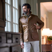 A man in a brown jacket and light pants stands indoors by a window, looking outside with one hand in his pocket.
