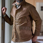 Man wearing a brown waxed jacket with large pockets, standing indoors near a window with one hand resting on the frame.