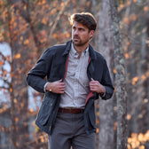 Man in a Tom Beckbe Piedmont Jacket over a checked shirt stands outdoors amid autumn forest foliage.