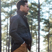 A man in a Tom Beckbe Piedmont Jacket and tan pants stands sideways in a forest with tall trees behind him.