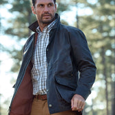 Man outdoors in a Tom Beckbe Piedmont Jacket and plaid shirt, standing in a wooded area with blurred trees behind.
