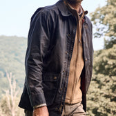 Man in a Tom Beckbe Piedmont Jacket over a beige sweater, strolling outdoors with trees and hills in the background.