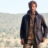 Man outdoors in a wooded landscape, wearing a Tom Beckbe Piedmont Jacket over a beige zip-up and shirt.