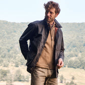 In a forested backdrop, someone in a Tom Beckbe Piedmont Jacket stands gazing sideways, hands in pockets.