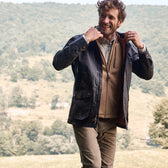 A man in a Tom Beckbe Piedmont Jacket and brown sweater stands in a grassy field with hills on a clear day.