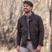 Man outdoors in a Tom Beckbe Piedmont Jacket over plaid shirt and khakis, smiling with trees behind him.