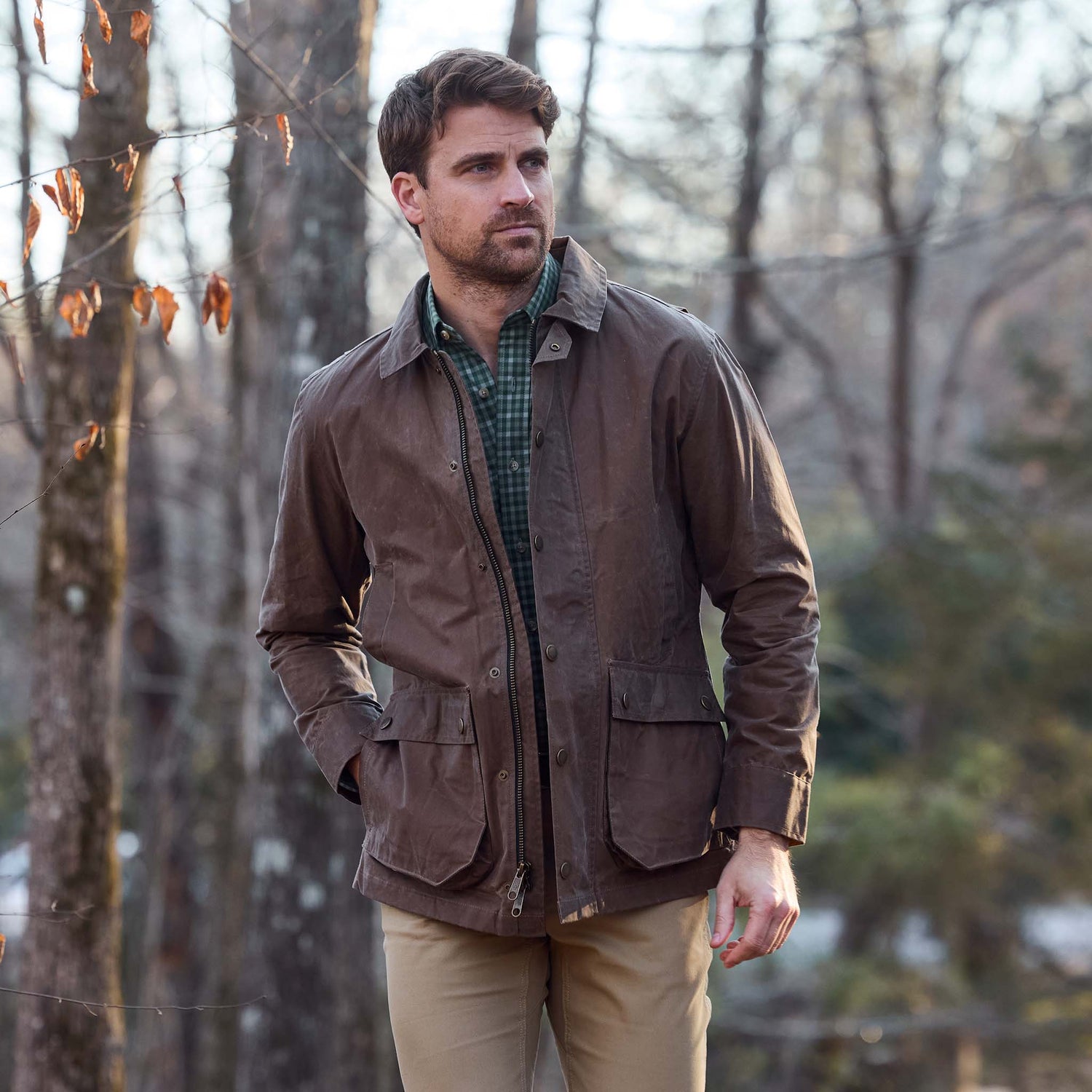 A man wears a Tom Beckbe Piedmont Jacket, green shirt, and tan pants while standing outdoors with hands in pockets.