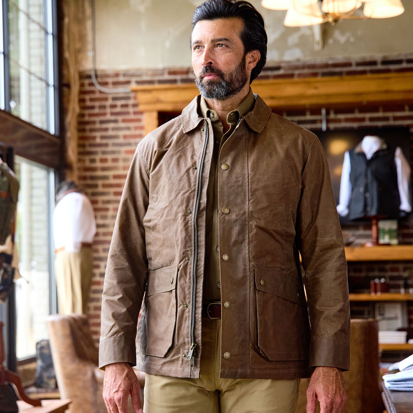 A man in a Tom Beckbe Piedmont Jacket and khaki pants stands indoors in a rustic room with brick walls and wood furniture.