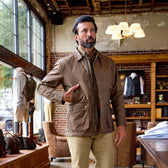 A man wears the Tom Beckbe Piedmont Jacket indoors near a desk, with clothing and bags displayed behind him.