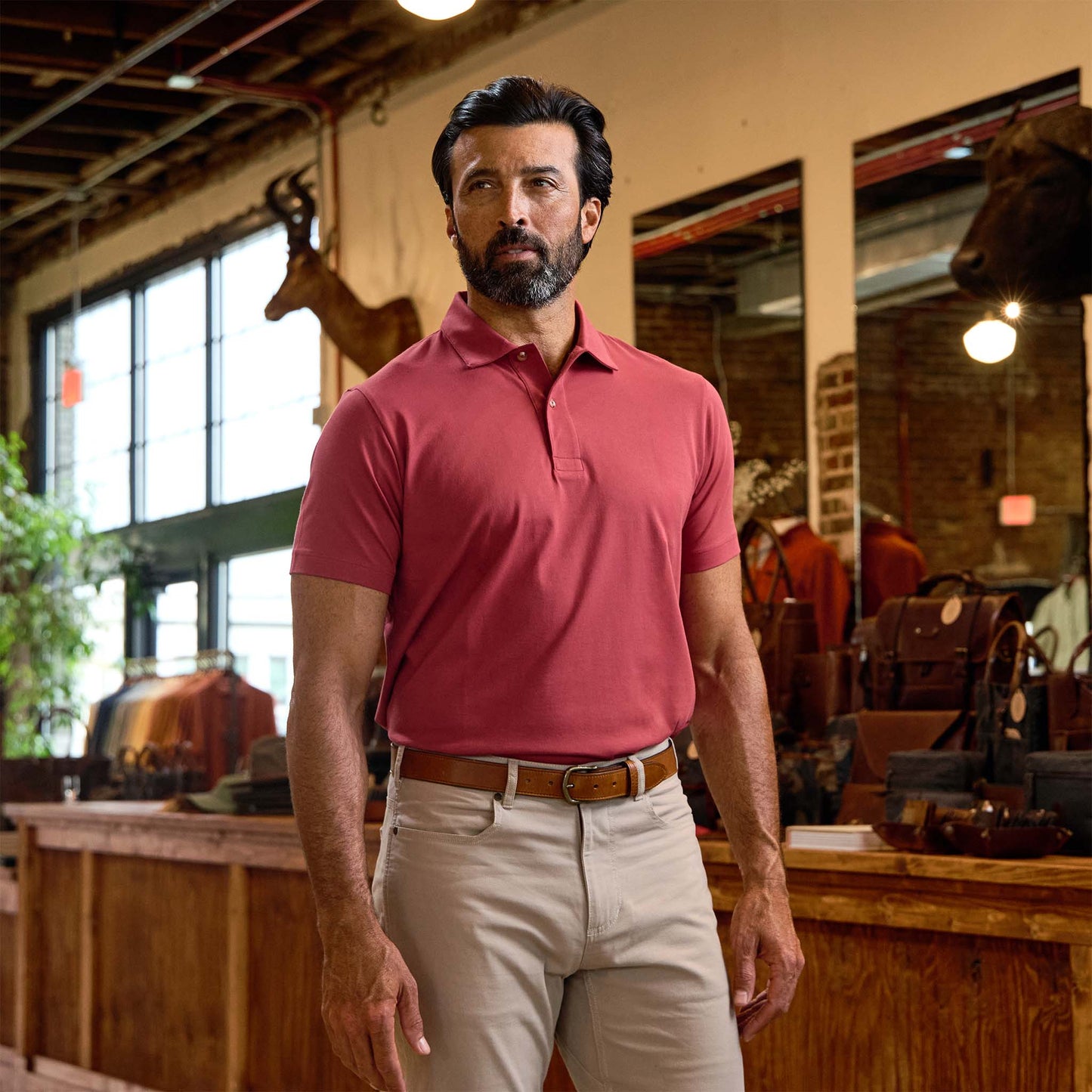 Man wearing a red polo shirt and beige pants stands inside a rustic store with leather goods and taxidermy on display.